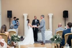 A bride and groom stand facing each other holding hands during a Bath Spa Hotel Weddings ceremony indoors, with guests seated and a photographer capturing the moment.