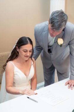 A bride in a white dress sits at a table, reviewing a document, while a man in a light gray suit with a boutonnière stands beside her, looking at the paper—capturing an elegant moment at Bath Spa Hotel Weddings.