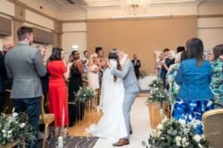 A bride and groom kiss at the end of the aisle while wedding guests stand, clap, and take photos in a beautifully decorated indoor venue at Bath Spa Hotel Weddings.