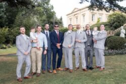 A group of men and two young children pose for a formal photo on a lawn in front of a large building, dressed in suits and ties—capturing the elegance often seen at Bath Spa Hotel Weddings.