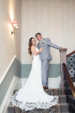 A bride in a white dress and a groom in a light gray suit pose together on a carpeted staircase indoors, capturing the elegance of Bath Spa Hotel Weddings.