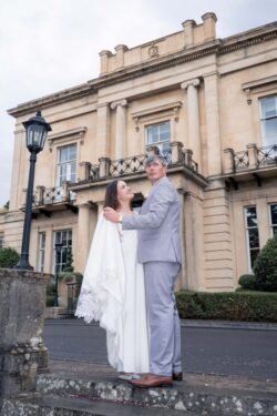 A bride and groom stand together outside a historic stone building at Bath Spa Hotel Weddings, looking up. The groom wears a light gray suit; the bride stuns in a white dress with a flowing cape.