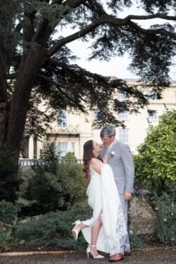 A bride and groom stand closely together outdoors under a large tree at Bath Spa Hotel Weddings, looking at each other and smiling, with a building and greenery in the background.