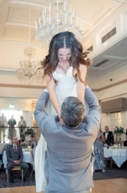 A man in a suit lifts a woman in a white dress at an indoor event, likely a Bath Spa Hotel Weddings reception, with seated guests and chandeliers glowing in the background.