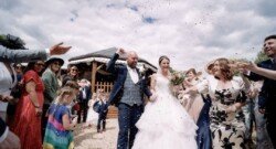 A bride and groom walk hand-in-hand at Orchardleigh Weddings as guests throw confetti. The bride wears a white gown, and the groom is in a blue suit. The background shows a wooden structure under a partly cloudy sky.