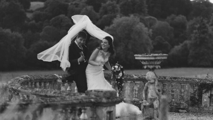 A bride and groom laugh as they walk together at Orchardleigh Weddings. The groom playfully holds the bride's veil over his head. They are outdoors on a stone terrace with a forested background.