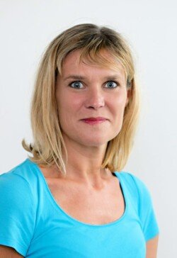 A woman with shoulder-length blonde hair wearing a light blue shirt poses against a plain white background, exemplifying the professional touch offered by our Corporate Photography & Video Services.