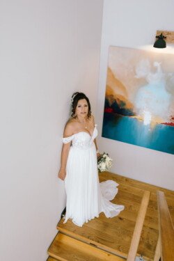 A bride in an off-the-shoulder wedding dress stands on a wooden staircase holding a bouquet, next to a colorful abstract painting, capturing the charm of Croftview Weddings.