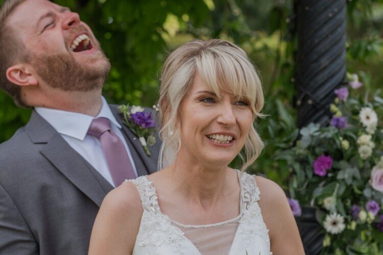 A bride and groom share a joyful moment outdoors; the bride smiles warmly while the groom laughs heartily. They stand in front of green foliage and a floral arrangement, beautifully captured by their weddings photographer at Elmhay Park.