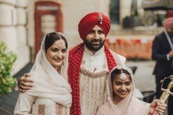A man wearing a red turban and traditional attire stands with two women dressed in traditional clothing, all smiling, captured beautifully by Fuji Cameras for weddings.