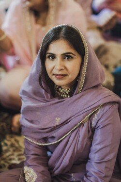A woman wearing a purple traditional outfit and headscarf, sitting indoors and looking towards the camera. The photo, taken with Fuji Cameras for weddings, captures her elegance and poise perfectly.