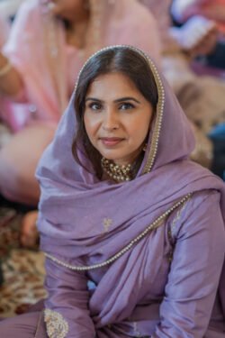 A woman in a lilac traditional outfit is sitting at the Guildhall Bath. She is wearing a matching headscarf, pearl necklace, and has a neutral expression. Other people can be seen in the blurred background.