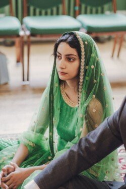 A woman wearing a green dress and matching embroidered headscarf sits pensively during an indoor event with green chairs in the background, capturing the essence of the moment as if through Fuji Cameras for weddings.