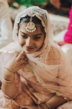 A young girl dressed in a pink traditional outfit with a jeweled headpiece and a sheer scarf smiles, sitting indoors—a perfect moment captured by Fuji Cameras for weddings.