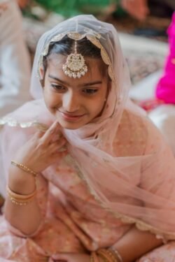 A young girl, wearing a light pink traditional outfit and headscarf adorned with a maang tikka, smiles gently. Seated indoors in the historic Guildhall Bath, she has gold bangles on her wrist, capturing a timeless elegance.