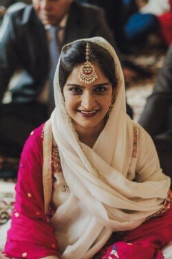 A woman wearing a white headscarf with a decorative headpiece and a vibrant pink outfit smiles while seated indoors. A man in a suit is partially visible in the background, capturing the scene's essence perfectly suited for Fuji Cameras for weddings.