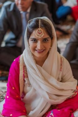 A woman wearing a white headscarf and a pink traditional outfit smiles at the camera in front of the historic Guildhall Bath. She is adorned with a decorative headpiece, and other people are visible in the background.