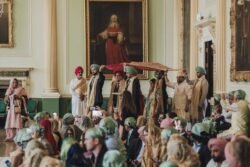 People dressed in traditional attire gather in the ornate Guildhall, with large portraits adorning the walls. Some hold umbrellas over those standing, while others sit, observing and capturing moments as a wedding photographer discreetly mingles among them.