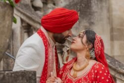 A couple dressed in traditional attire, with the man wearing a red turban and the woman in a red gown with gold jewelry, gazing at each other outdoors near stone steps leading to the Guildhall Bath.