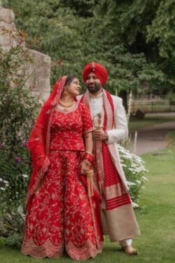 A bride and groom in traditional red and white attire stand in a garden at the Guildhall Bath, surrounded by greenery and flowers.