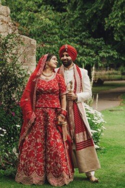 A couple wearing traditional Indian wedding attire stands together in a garden; the woman in a red lehenga and the man in a white sherwani with a matching red turban, expertly captured using Fuji Cameras for weddings.