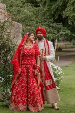 A couple dressed in traditional red and white South Asian attire stand outside in a garden by the Guildhall, smiling at each other.