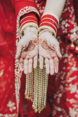 Close-up of hands with intricate henna designs and the initials "RS," adorned with gold jewelry and red, embroidered sleeves captured perfectly by Fuji Cameras for weddings.