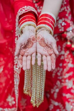 Close-up of a person wearing red and gold bangles, displaying intricate henna designs on their open palms. The person is dressed in red fabric with detailed embellishments, reminiscent of the opulent decor found at Guildhall Bath.