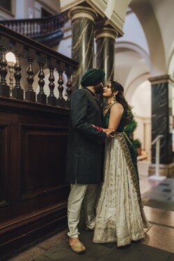 A couple stands in an ornate hallway. The man wears a dark sherwani and a green turban, while the woman dons a white and green traditional dress. They stand close, gazing into each other's eyes, their moment beautifully captured by Fuji Cameras for weddings.