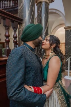 A couple dressed in traditional South Asian attire stands close together, gazing into each other's eyes, next to an ornate wooden and marble staircase in the grand Guildhall Bath.
