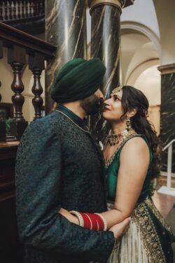 A couple in traditional attire stands closely, facing each other. The man wears a green turban and embroidered outfit, while the woman is in a green and white dress with jewelry. Marble columns are in the background, capturing a moment beautifully suited for any Guildhall Bath Wedding Photographer's portfolio.