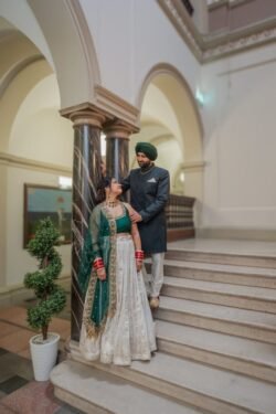 A couple stands on the stairs of the Guildhall, with the man in a green turban and gray outfit and the woman in a green and white traditional dress. They are looking at each other, standing by ornate columns bathed in soft light.