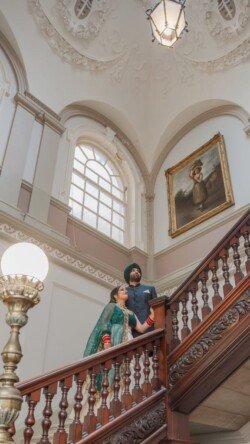 A man and woman in traditional attire stand on a grand wooden staircase in an ornate room with a high ceiling, large window, and a painting on the wall above the staircase, capturing the timeless elegance of the Guildhall.