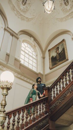 A couple stands on a wooden staircase inside a grand, ornately decorated building with white walls and a large window, capturing the magic of the moment with their Fuji Cameras for weddings.