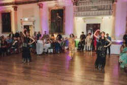 Three women in black dresses perform a dance in a grand hall with an audience seated at tables. Portraits adorn the walls, and colorful lights illuminate the scene, perfect for capturing with Fuji Cameras for weddings.