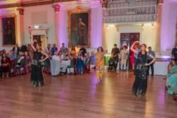 Three dancers perform in traditional attire on a wooden floor in the beautifully decorated Bath Guildhall. The audience sits entranced, surrounded by historical paintings, ornate walls, and soft uplighting that enhances the hall's timeless charm.