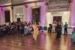 A group of women in traditional attire dance in a decorated hall with seated guests watching, capturing every vibrant moment with Fuji Cameras for weddings. Large portraits and ornate decorations are visible on the walls.