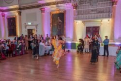A group of people dressed in traditional attire performs a dance in the beautifully decorated Guildhall Bath, with seated guests watching in admiration.