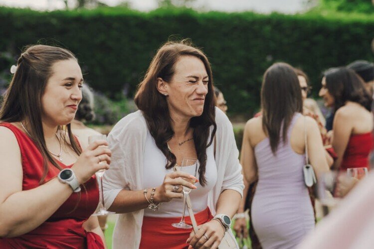 Two women holding glasses make expressive faces while socializing at an outdoor event, perhaps captured by a Farleigh House Wedding Photographer. One woman wears a white sweater over a red dress, and the other is also in a red dress. Other attendees mingle in the background.