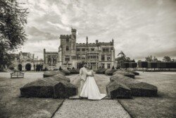 A couple in formal attire stands in front of Farleigh House, a large historic castle with groomed hedges and a dramatic cloudy sky, captured beautifully by their wedding photographer.
