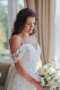 A woman in a white off-the-shoulder wedding dress holding a bouquet of white and green flowers stands by a window with beige curtains at Orchardleigh House. She has long dark hair and gazes down at the bouquet.