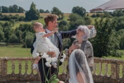 A man holding a child adjusts a woman's hat while another person in a wedding dress is seated nearby. They are outdoors at Orchardleigh House, with a scenic view of a grassy landscape and trees in the background.