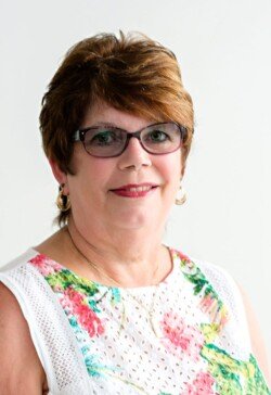 A woman with short brown hair, glasses, and hoop earrings is wearing a white sleeveless top with a floral pattern. She is smiling and looking at the camera against a plain background, perfect for corporate photography & video services.