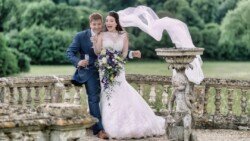 A bride and groom stand outdoors by a stone railing at Orchardleigh Weddings, with the groom holding a champagne flute and the bride holding a bouquet. The bride's veil is blown by the wind.