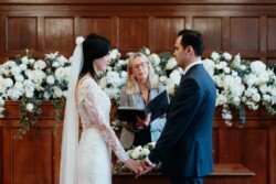 A bride and groom stand holding hands during a beautiful Guildhall Bath Weddings ceremony officiated by a woman. They are facing each other in front of a wooden wall adorned with white flowers, creating an enchanting atmosphere.