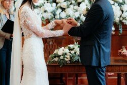 A couple exchanges rings during a Guildhall Bath Weddings ceremony, with the bride in a lace dress and the groom in a dark suit. A floral arrangement and officiant are visible in the background.