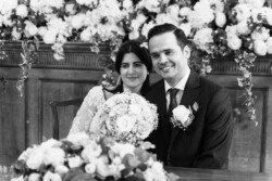 A couple in formal attire poses for a wedding photo at the Guildhall Bath Weddings, sitting in front of a floral arrangement with bouquets in the foreground.