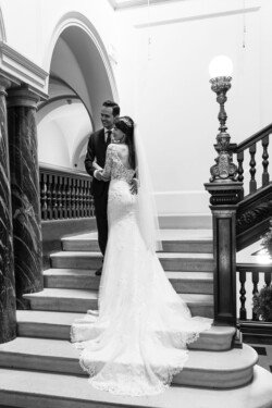 A bride and groom stand on a staircase at Guildhall Bath Weddings, with the bride in a long white gown and veil and the groom in a dark suit. They share a tender moment while posing for a photograph.