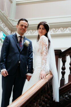 A couple stands together on a staircase, dressed in wedding attire at their Guildhall Bath wedding. The groom wears a dark suit and tie, while the bride is in a white lace gown with a veil, holding the staircase railing.