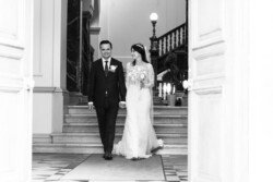 A bride and groom walk hand in hand down the steps of Guildhall Bath, both smiling. The image is black and white, capturing the timeless elegance of their wedding day. The bride is in a stunning gown while the groom wears a classic suit.